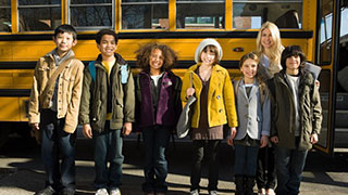 Image of six young students and one adult standing along side a school bus, smiling.