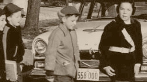 Black and white archive photo of a student Patroller, wearing a patrol belt, helping kids cross the road.